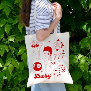 Puede incluir: Bolso tote color crema con ilustraciones rojas. El diseño incluye un cóctel, una rosa, el número 777, un bebé diablo, una bola ocho, dados y la palabra "Lucky". El bolso lo lleva una persona con una camisa a rayas.