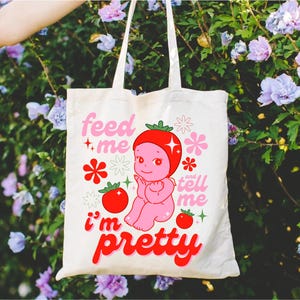 May include: A white tote bag with a pink and red cartoon character on it. The character is a baby with a strawberry on its head. The text on the bag says "Feed me and tell me I'm pretty".