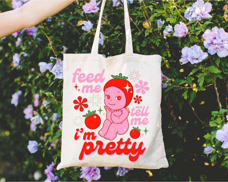 May include: A white tote bag with a pink and red cartoon character on it. The character is a baby with a strawberry on its head. The text on the bag says "Feed me and tell me I'm pretty".