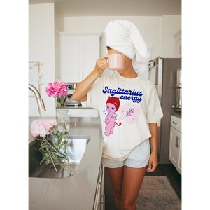Puede incluir: Una camiseta blanca con una ilustraci&oacute;n de dibujos animados de un diablo rosa con cuernos rojos y un s&iacute;mbolo de Sagitario azul y blanco. El texto "Sagittarius energy" est&aacute; impreso en azul en la camiseta.