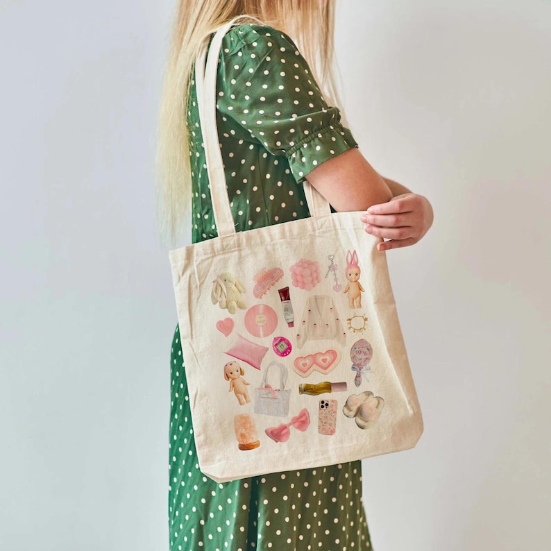 May include: A white canvas tote bag with a pink and white design featuring various items like a doll, a heart, a lipstick, and a phone. The bag is being held by a person wearing a green polka dot dress.