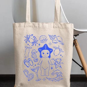 May include: Beige canvas tote bag with blue illustrations of beach-themed elements. The design includes a starfish-headed figure, seashells, a crab, a seahorse, a cocktail, and other summer-related graphics. The bag has two long handles.