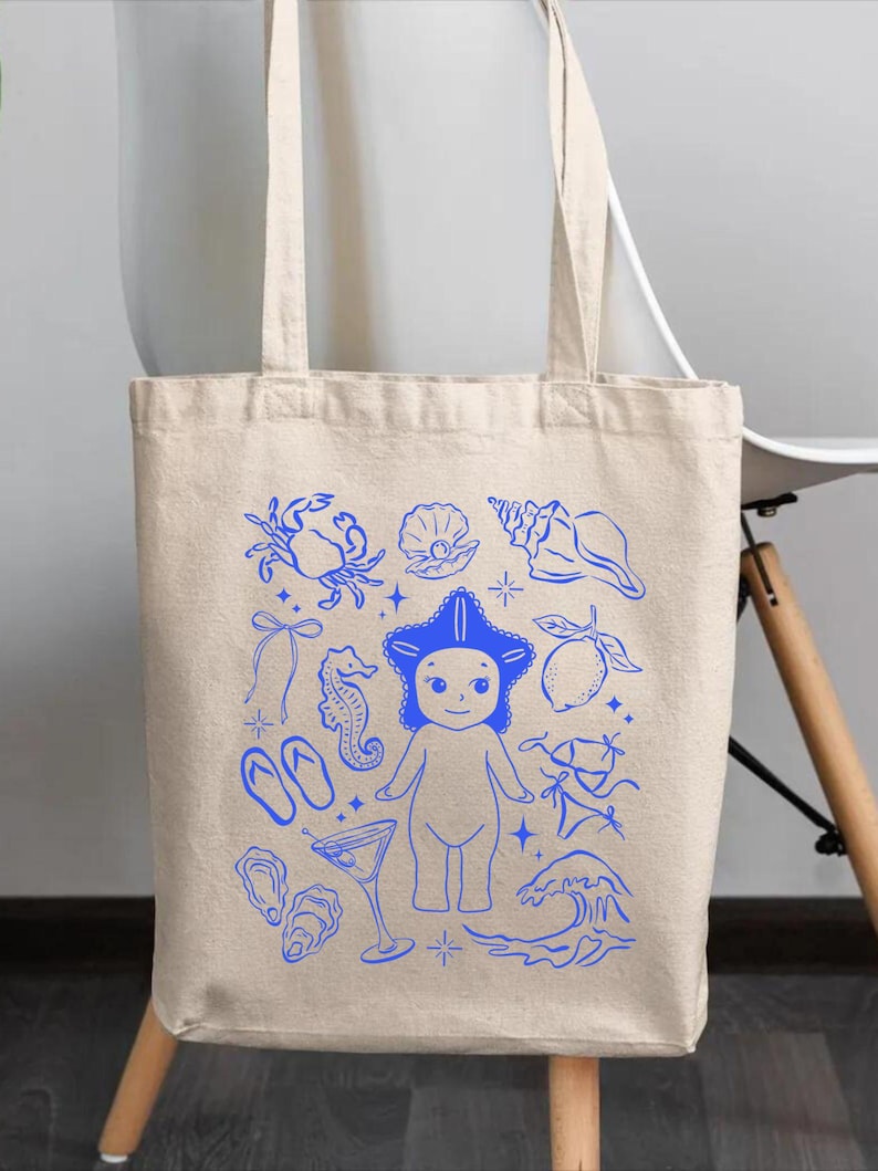 May include: Beige canvas tote bag with blue illustrations of beach-themed elements. The design includes a starfish-headed figure, seashells, a crab, a seahorse, a cocktail, and other summer-related graphics. The bag has two long handles.