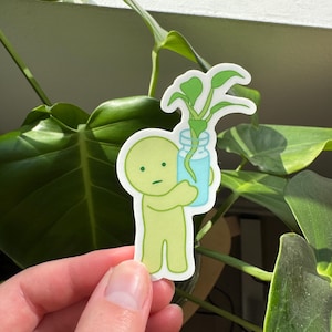 May include: A green cartoon character holding a glass jar with a small green plant inside. The character has a simple, round face with a single eye and a small mouth.