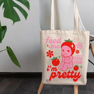 May include: A white canvas tote bag with a pink and red illustration of a cartoon character with a tomato on its head. The text on the bag reads "Feed me, tell me I'm pretty".