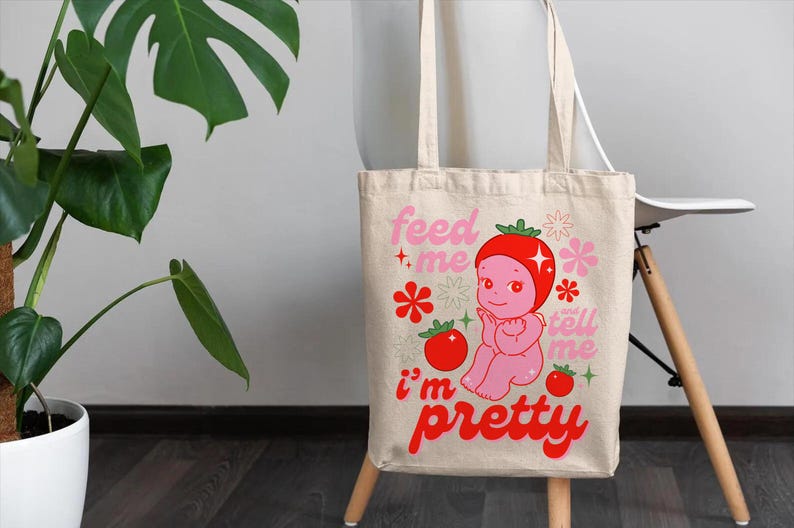 May include: A white canvas tote bag with a pink and red illustration of a cartoon character with a tomato on its head. The text on the bag reads "Feed me, tell me I'm pretty".