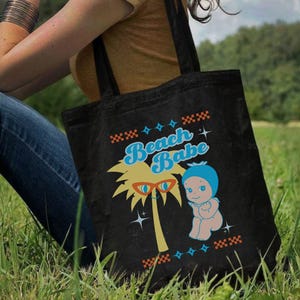 May include: Black tote bag with a colorful graphic that says "Beach Babe". The graphic includes a palm tree with sunglasses, a baby in a blue bonnet, and decorative star and cross patterns. The bag has two black handles.