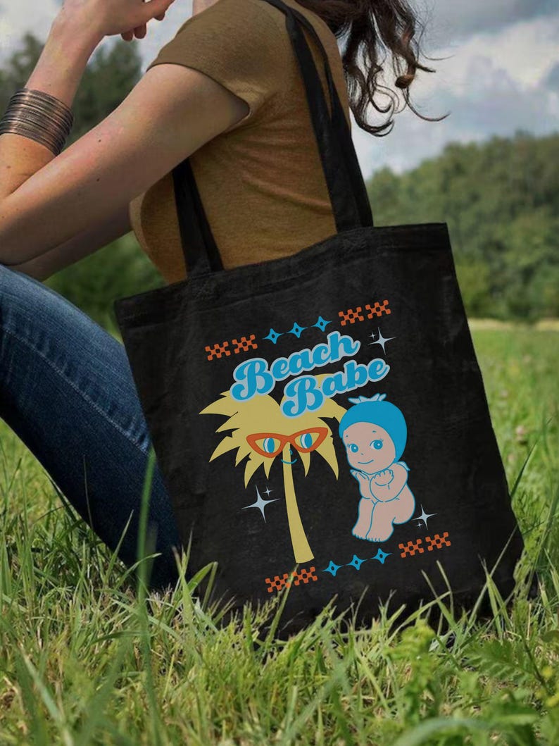 May include: Black tote bag with a colorful graphic that says "Beach Babe". The graphic includes a palm tree with sunglasses, a baby in a blue bonnet, and decorative star and cross patterns. The bag has two black handles.