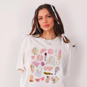 May include: A white t-shirt with a pink and white graphic print of various items, including a heart, a bunny, a hair clip, a phone case, a bag, and a pair of slippers.