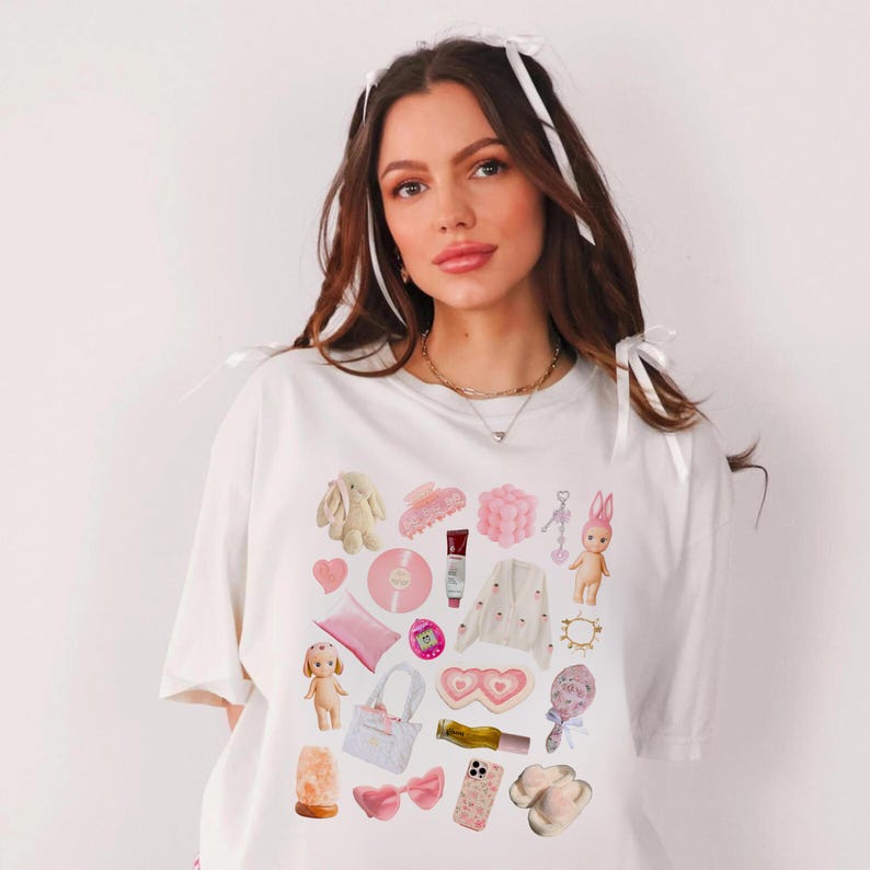 May include: A white t-shirt with a pink and white graphic print of various items, including a heart, a bunny, a hair clip, a phone case, a bag, and a pair of slippers.