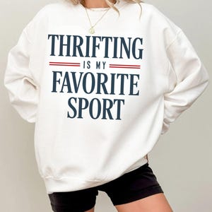 May include: White sweatshirt with the text "THRIFTING IS MY FAVORITE SPORT" in navy blue lettering with red stripes. The sweatshirt is a crew neck style.
