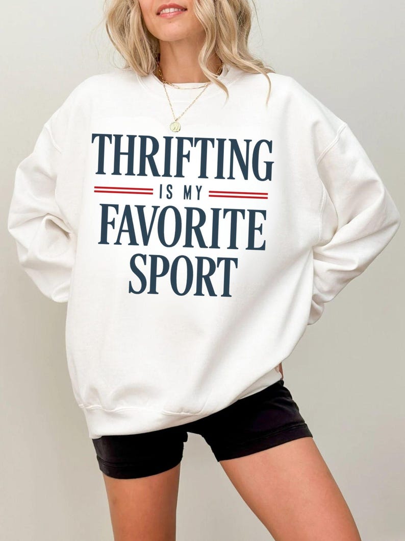 May include: White sweatshirt with the text "THRIFTING IS MY FAVORITE SPORT" in navy blue lettering with red stripes. The sweatshirt is a crew neck style.