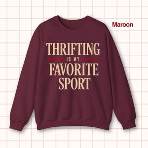 May include: Maroon crewneck sweatshirt with the text "Thrifting is my favorite sport" in a cream-colored font. The text is centered on the front of the sweatshirt, with two red lines separating the phrases. The sweatshirt is a solid maroon color.