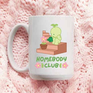 May include: White ceramic mug featuring a cartoon illustration of a green character watering a potted flower on brown steps. The mug has the text "HOMEBODY CLUB" in green and pink. The mug has a large handle.