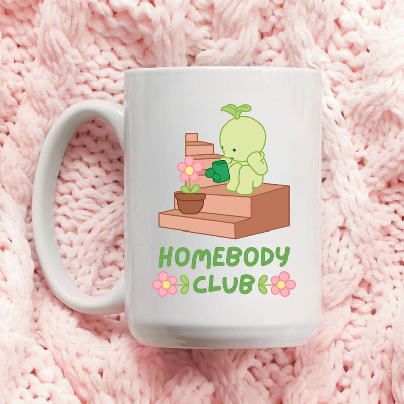 May include: White ceramic mug featuring a cartoon illustration of a green character watering a potted flower on brown steps. The mug has the text "HOMEBODY CLUB" in green and pink. The mug has a large handle.