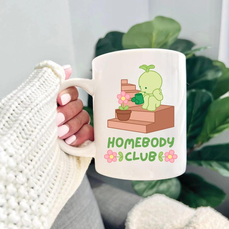 May include: White ceramic mug featuring a cartoon illustration of a green character watering a flower on a set of stairs. The mug has the text "HOMEBODY CLUB" with flower accents. The mug is held by a person wearing a white sweater.