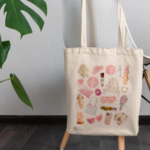 May include: A white canvas tote bag with a pink and white design. The design features a variety of items, including a doll, a heart, a comb, a bottle of nail polish, a pair of sunglasses, and a pair of earrings.