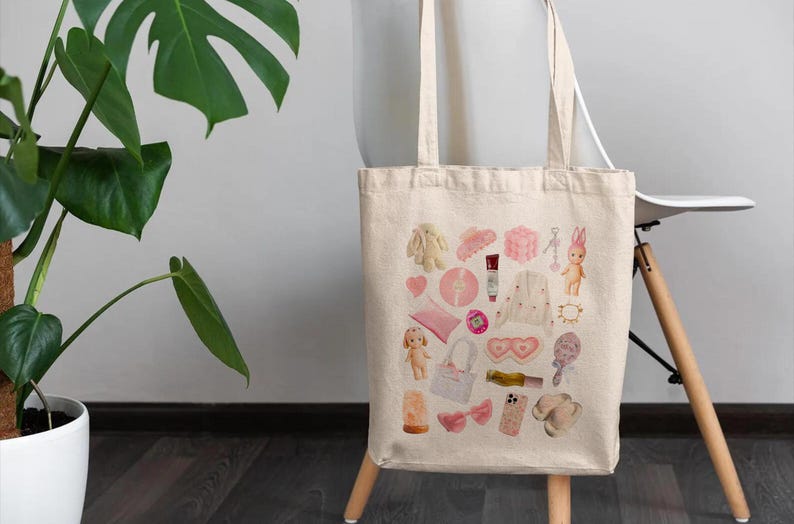 May include: A white canvas tote bag with a pink and white design. The design features a variety of items, including a doll, a heart, a comb, a bottle of nail polish, a pair of sunglasses, and a pair of earrings.