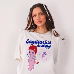 Puede incluir: Camiseta blanca con un dibujo animado de diablo rojo y azul con un halo rojo y una cola azul. El texto "Sagittarius energy" est&aacute; impreso en azul en la camiseta. Una nube rosa con un s&iacute;mbolo de Sagitario azul est&aacute; detr&aacute;s del diablo.
