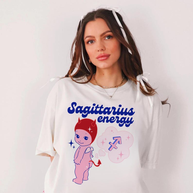 Puede incluir: Camiseta blanca con un dibujo animado de diablo rojo y azul con un halo rojo y una cola azul. El texto "Sagittarius energy" est&aacute; impreso en azul en la camiseta. Una nube rosa con un s&iacute;mbolo de Sagitario azul est&aacute; detr&aacute;s del diablo.