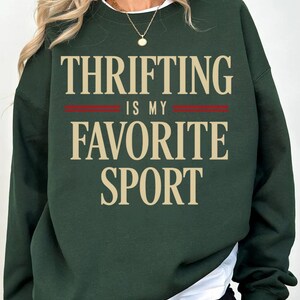 May include: Dark green sweatshirt with the text "THRIFTING IS MY FAVORITE SPORT" in a beige font. The text is centered on the front of the sweatshirt, with a red stripe separating the lines. The sweatshirt has a white crew neck collar.