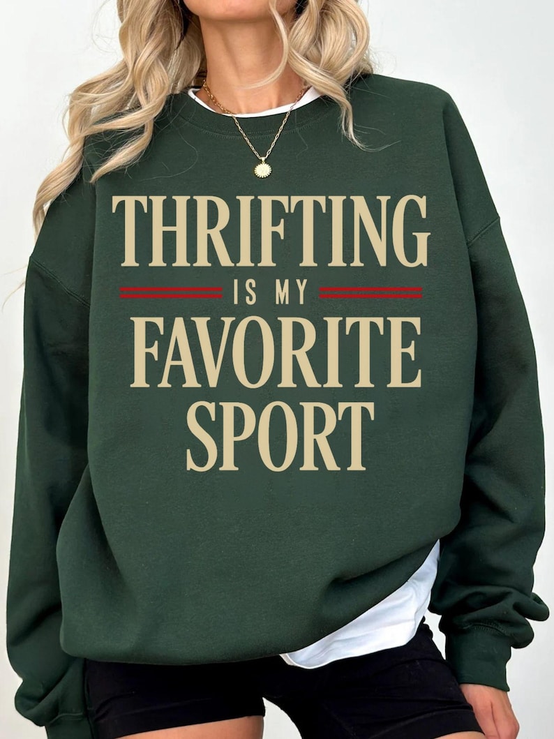 May include: Dark green sweatshirt with the text "THRIFTING IS MY FAVORITE SPORT" in a beige font. The text is centered on the front of the sweatshirt, with a red stripe separating the lines. The sweatshirt has a white crew neck collar.