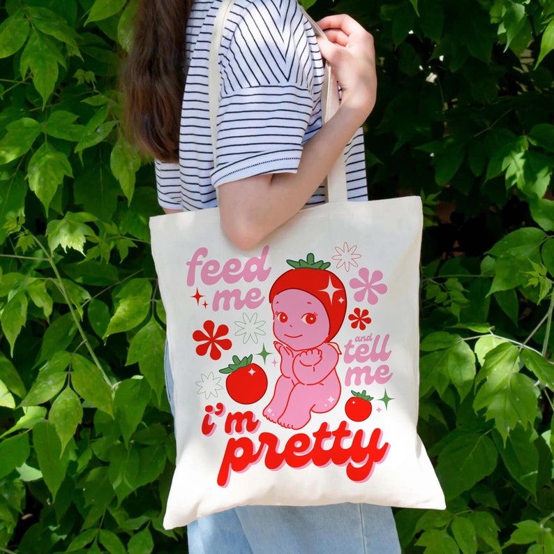 May include: A white tote bag with a pink and red cartoon strawberry character. The text on the bag reads "feed me and tell me i'm pretty".