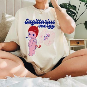 Puede incluir: Camiseta blanca con un dibujo animado de un diablo con cuernos rojos y un cuerpo rosa. La camiseta tiene el texto "Sagittarius energy" en letras azules y un s&iacute;mbolo de Sagitario rosa en una nube.