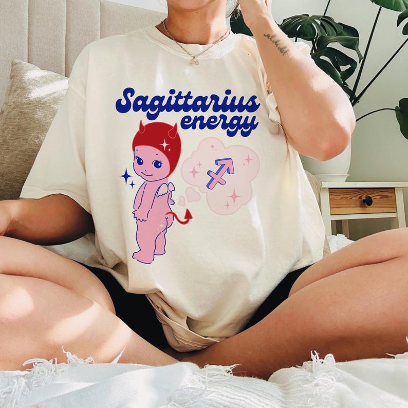 Puede incluir: Camiseta blanca con un dibujo animado de un diablo con cuernos rojos y un cuerpo rosa. La camiseta tiene el texto "Sagittarius energy" en letras azules y un s&iacute;mbolo de Sagitario rosa en una nube.