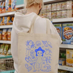 May include: Beige tote bag with blue illustrations of sea creatures and beach-themed elements. The design includes a starfish-headed figure, seashells, a crab, seahorse, and waves. The bag has two beige straps and is carried over a person's shoulder.