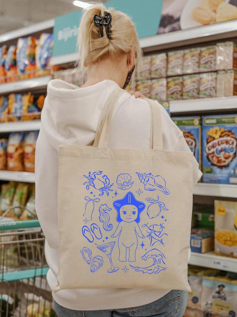 May include: Beige tote bag with blue illustrations of sea creatures and beach-themed elements. The design includes a starfish-headed figure, seashells, a crab, seahorse, and waves. The bag has two beige straps and is carried over a person's shoulder.