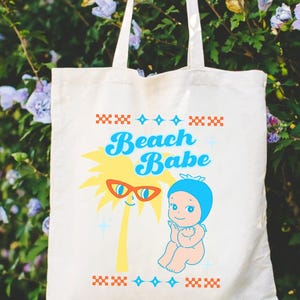 May include: Cream-colored tote bag with the words "Beach Babe" in blue script. The bag features a cartoon palm tree wearing sunglasses and a baby in a blue bonnet. The design includes red and blue decorative elements.