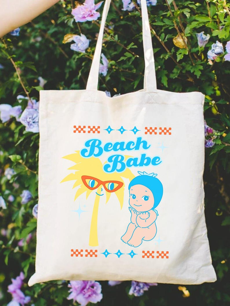 May include: Cream-colored tote bag with the words "Beach Babe" in blue script. The bag features a cartoon palm tree wearing sunglasses and a baby in a blue bonnet. The design includes red and blue decorative elements.