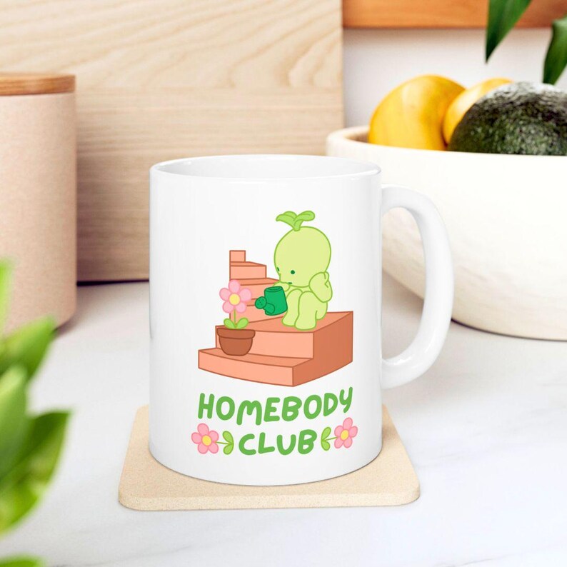 May include: White ceramic mug featuring a cartoon illustration of a green creature watering a flower on a set of stairs. The mug has the text "HOMEBODY CLUB" with flower accents. The mug is on a beige coaster.