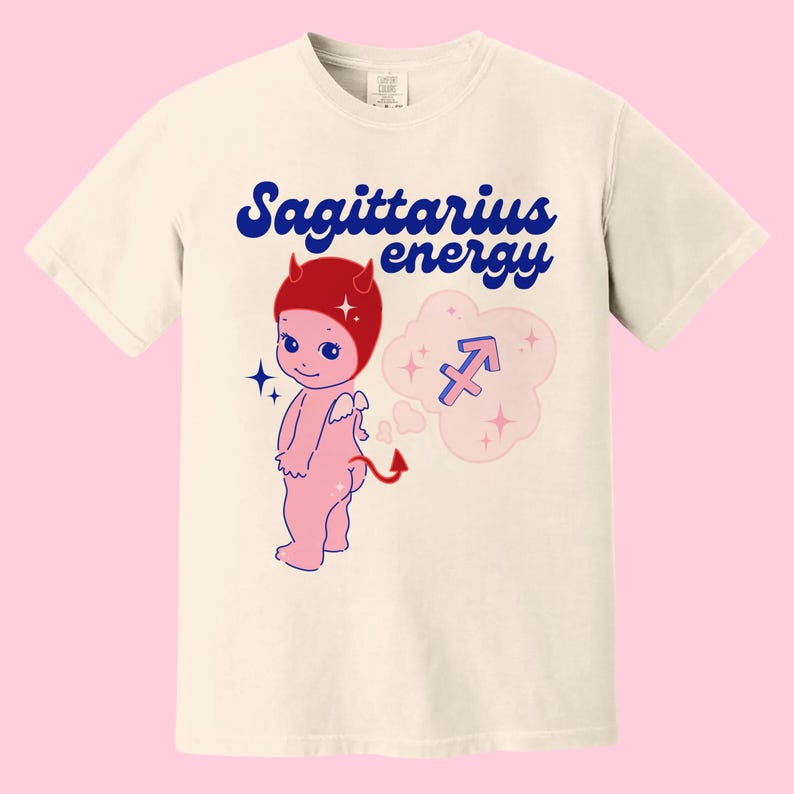 Puede incluir: Una camiseta blanca con una imagen de dibujos animados de un beb&eacute; diablo rosa con cuernos rojos y un gorro rojo. El beb&eacute; est&aacute; sosteniendo su cola y tiene una nube detr&aacute;s con el s&iacute;mbolo del zod&iacute;aco Sagitario y el texto "Energ&iacute;a Sagitario".
