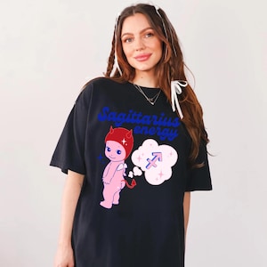Puede incluir: Camiseta negra con un diablo de dibujos animados rosa con cuernos rojos y un halo azul. El diablo sostiene una cola y tiene una burbuja de pensamiento sobre su cabeza con un s&iacute;mbolo de Sagitario azul y estrellas blancas. El texto "Sagittarius energy" est&aacute; impreso en azul en la camiseta.