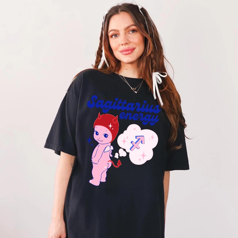 Puede incluir: Camiseta negra con un diablo de dibujos animados rosa con cuernos rojos y un halo azul. El diablo sostiene una cola y tiene una burbuja de pensamiento sobre su cabeza con un s&iacute;mbolo de Sagitario azul y estrellas blancas. El texto "Sagittarius energy" est&aacute; impreso en azul en la camiseta.