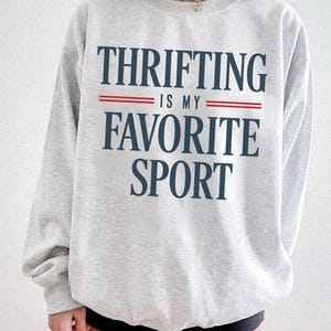 May include: A light gray sweatshirt with the text "THRIFTING IS MY FAVORITE SPORT" in navy blue, with red lines separating the phrases. The sweatshirt has long sleeves and a crew neck.