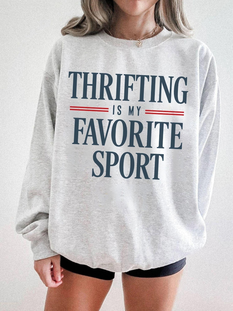 May include: A light gray sweatshirt with the text "THRIFTING IS MY FAVORITE SPORT" in navy blue, with red lines separating the phrases. The sweatshirt has long sleeves and a crew neck.