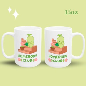 May include: Two white ceramic mugs with a whimsical design. Each mug features a green cartoon character watering a flower on a brown structure, with the text "HOMEBODY CLUB" and a flower graphic. The mugs are labeled "15oz".