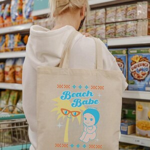 May include: Beige tote bag with the words "Beach Babe" in turquoise and a cartoon palm tree wearing sunglasses. A cartoon baby in a blue bonnet is also featured. The bag has tan straps and is being carried by a person.