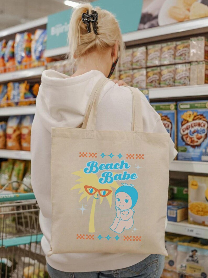 May include: Beige tote bag with the words "Beach Babe" in turquoise and a cartoon palm tree wearing sunglasses. A cartoon baby in a blue bonnet is also featured. The bag has tan straps and is being carried by a person.