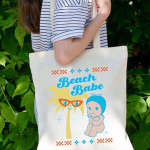 May include: Cream-colored tote bag featuring a retro design with the words "Beach Babe" in blue script. The design includes a palm tree wearing sunglasses and a baby figure. The bag is held by a person wearing a striped shirt.