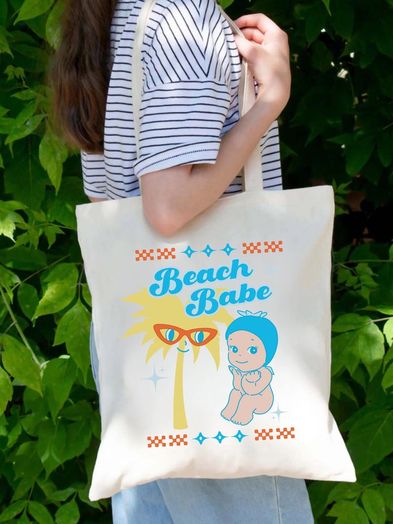 May include: Cream-colored tote bag featuring a retro design with the words "Beach Babe" in blue script. The design includes a palm tree wearing sunglasses and a baby figure. The bag is held by a person wearing a striped shirt.