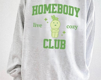 May include: A light gray sweatshirt with the words "HOMEBODY CLUB" in green block letters. The design includes a cartoon figure and the words "live" and "cozy" in green. The sweatshirt is a casual, comfortable style.