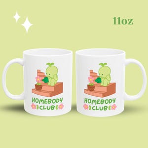 May include: Two white ceramic mugs with a whimsical illustration of a green character watering a flower on a set of steps. The mugs have the text "HOMEBODY CLUB" and are 11oz.