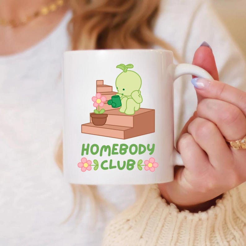 May include: White ceramic mug featuring a cartoon illustration of a green sprout watering a flower on a set of stairs. The mug has the text "HOMEBODY CLUB" with pink flower accents.