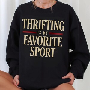 May include: Black sweatshirt with the text "THRIFTING IS MY FAVORITE SPORT" in a serif font. The text is in a light tan color with two red lines separating the phrases. The sweatshirt is worn by a person.