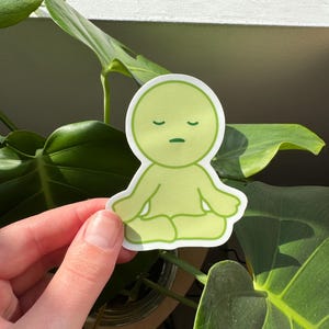 May include: A green sticker featuring a cartoon character in a meditative pose with closed eyes. The character has a simple, rounded design and is outlined in dark green.