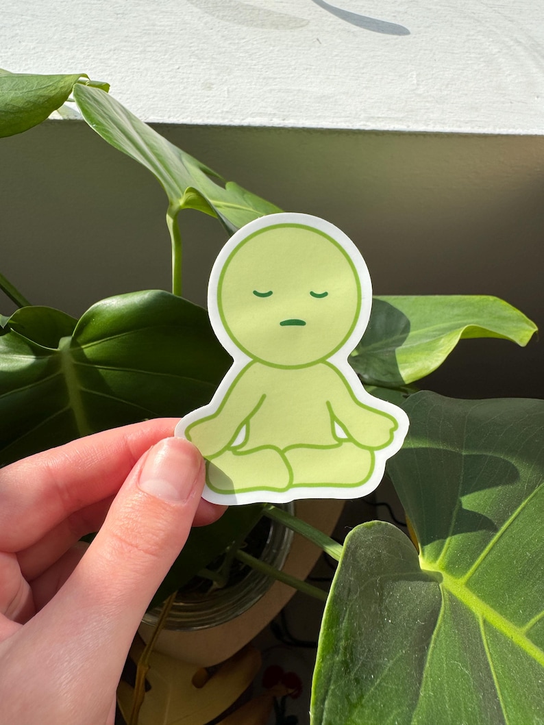 May include: A green sticker featuring a cartoon character in a meditative pose with closed eyes. The character has a simple, rounded design and is outlined in dark green.